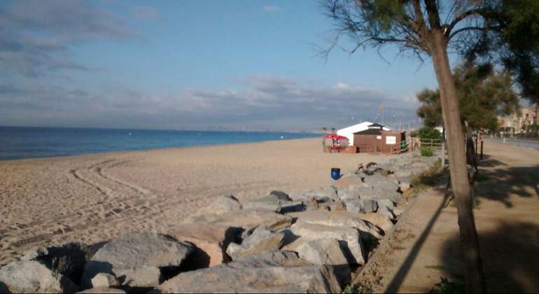 Chiringuito de playa en la arena de la zona de Ocata en Barcelona. Hay árboles en el paseo marítimo y marcas de neumáticos en la arena
