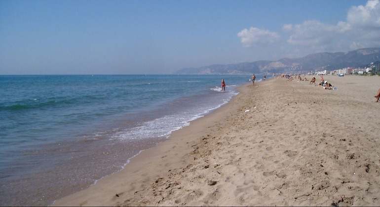 Playa de arena blanca con un pequeño salto al mar. Al fondo hay bañistas, pero no está muy llena la playa