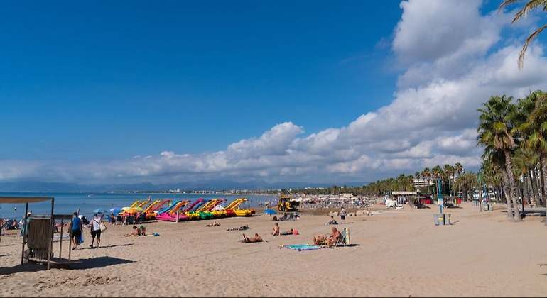 zona de arena con poca gente, se puede ver antes de entrar al mar una zona de alquiler de patines de agua de muchos colores y en el paseo marítimo una extensa zona de palmeras
