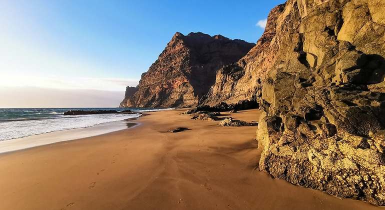 Una playa de arena marrón y agua transparente y calmada. Junto a la arena hay formaciones rocosas.