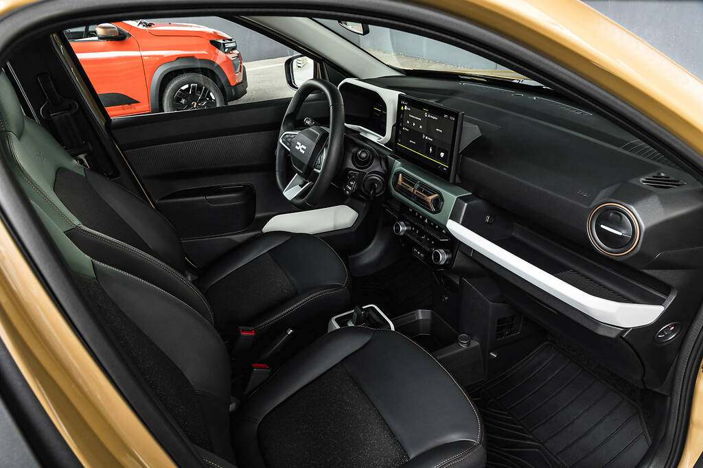 Dacia Spring 2024. Asientos (sin regulación de altura) de la versión anterior. El modelo urbano cuenta con una serie de accesorios que se pueden anclar a distintas partes del salpicadero, y que Dacia llama YouClip (por ejemplo, una linterna, un posavasos, un pequeño cajón y un soporte de móvil
) De serie llega con tres puntos de fijación, uno en el salpicadero y dos en la consola central.