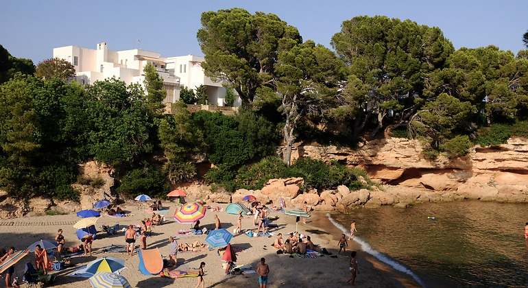 Personas y sombrillas en la playa con un pequeño peñón y árboles en el fondo tras los que resaltan unos edificios blancos